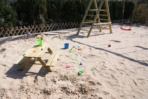 A sandy outdoor area with a wooden picnic table, a ladder, and a swing set. - Holiday home with 2 bedrooms (Timmendorfer Strand)
