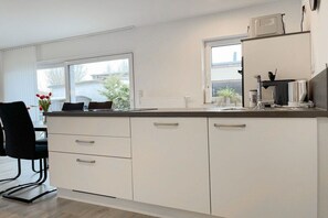 A modern kitchen with white cabinetry, a dining area with a black chair, and a window providing a view of the outside. - Holiday home with 2 bedrooms (Timmendorfer Strand)