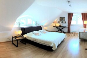 Ein Schlafzimmer mit einem Bett, Nachttischen, Lampen, einem kleinen Tisch und einem Fenster mit Vorhängen.