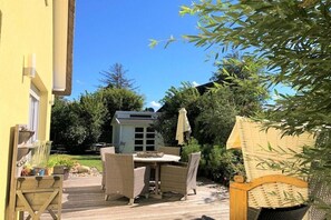 Ein Terrassenbereich im Freien mit einem runden Tisch und Korbstühlen, umgeben von viel Grün und blauem Himmel.