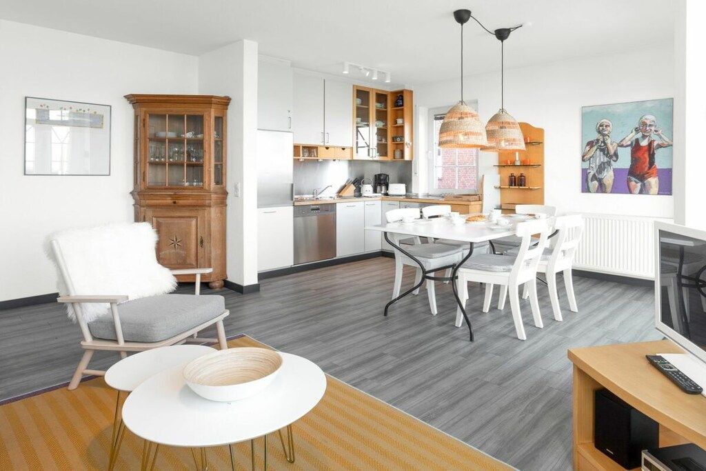 A modern kitchen with white cabinets, a dining area with a white table and chairs, and a living area with a TV and a sofa.