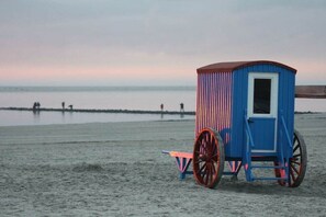 Una cabaña de playa con techo rojo y paredes azules, ubicada en una playa arenosa con el mar al fondo.