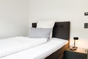 A neatly made bed with white bedding and striped pillows, a dark headboard, a bedside table with a lamp, and a small drawer. - Holiday home with 4 bedrooms (Timmendorfer Strand)
