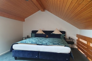 Ein Schlafzimmer mit schräg versetzter Decke, einem Bett mit blauer Bettdecke, zwei Nachttischen und einem hölzernen Kopfteil.