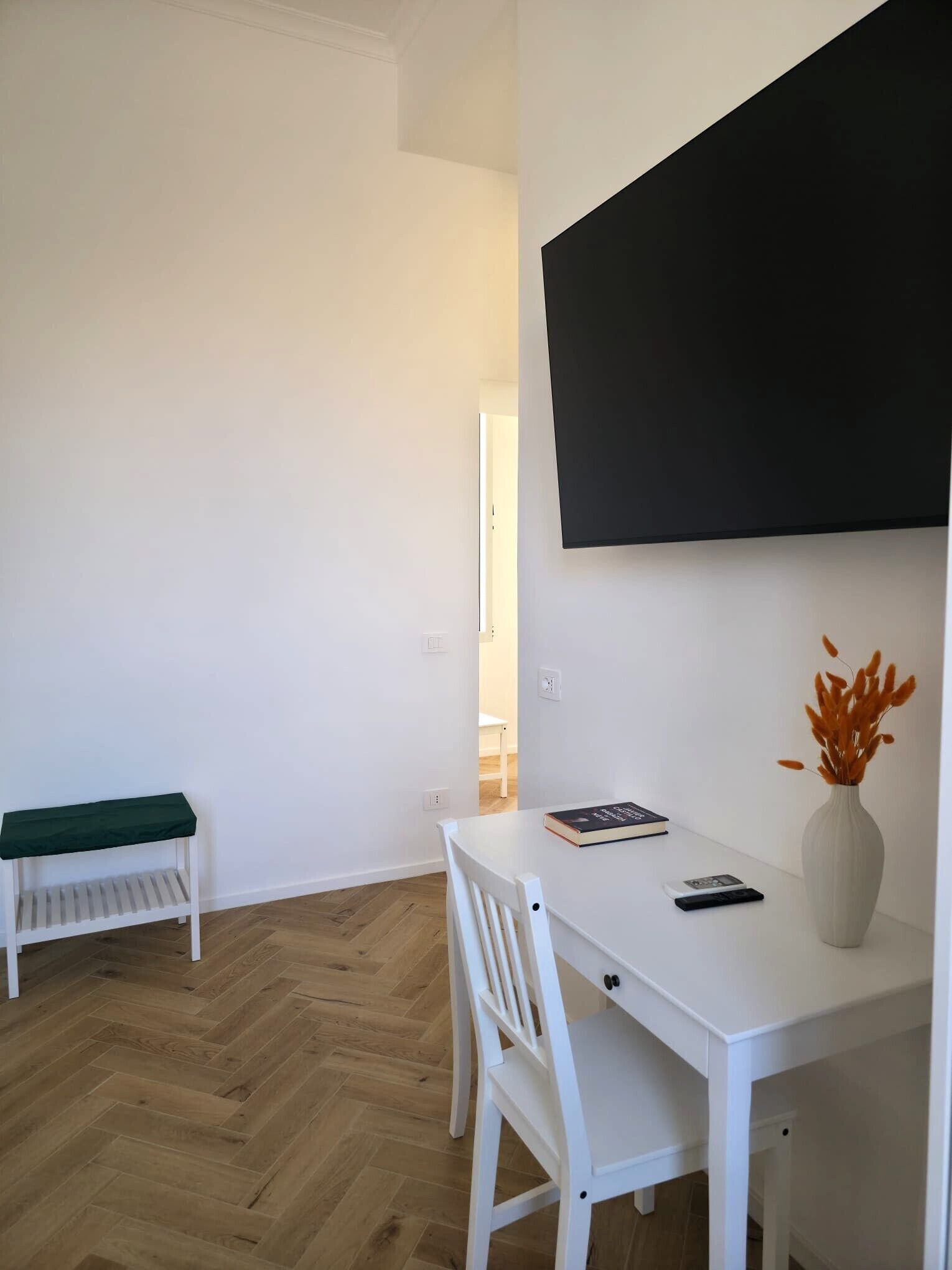 Et hvidt skrivebord med stol, et sort tv ophængt på væggen, en hvid bænk og en vase med orange blomster.