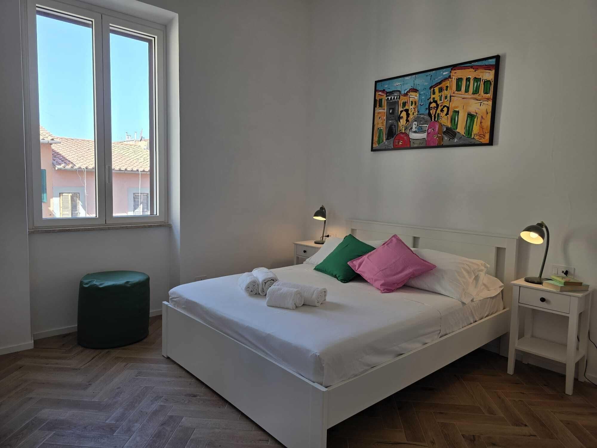 A hotel room with a bed, two bedside lamps, a painting on the wall, and a window with a view of buildings.