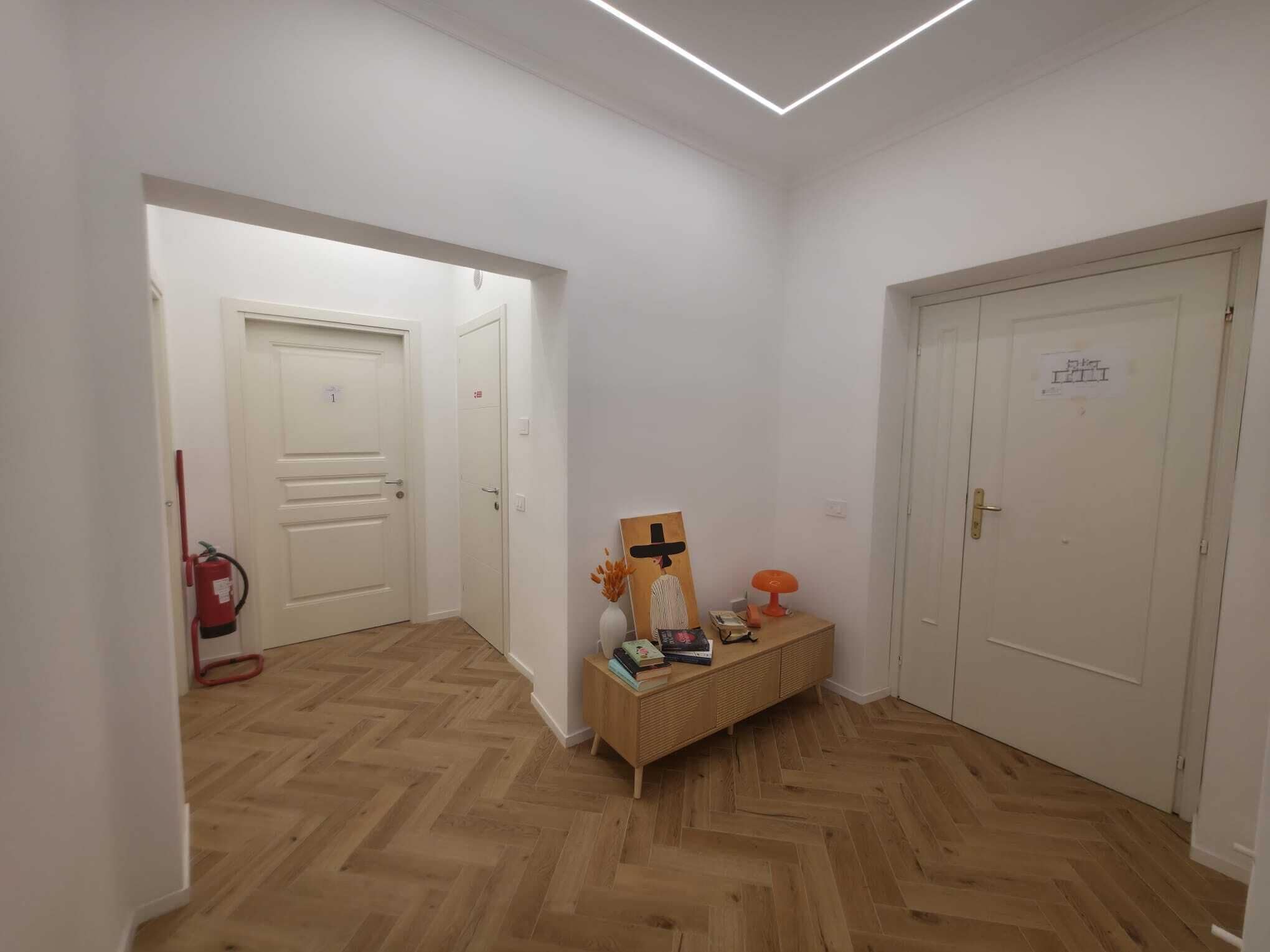 Un pasillo con suelo de madera, puertas blancas y una pequeña mesa con libros y una lámpara.