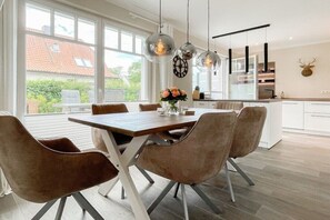 A modern dining area with a wooden table and cushioned chairs, large windows, and a kitchen in the background. - Holiday home with 3 bedrooms (Timmendorfer Strand)