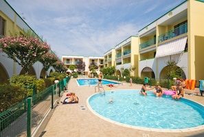 A swimming pool area with multiple pools, a fenced walkway, and a building with balconies. - Cozy flat with pool access (Bibione)