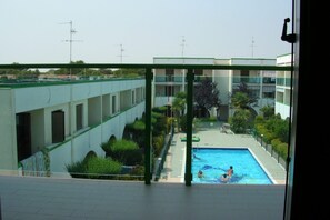 A view of a swimming pool with people swimming, surrounded by greenery and buildings with multiple antennas. - Cozy flat with pool access (Bibione)