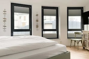 Ein Schlafzimmer mit einem Bett, einem Sessel und drei Fenstern mit schwarzen Rahmen.