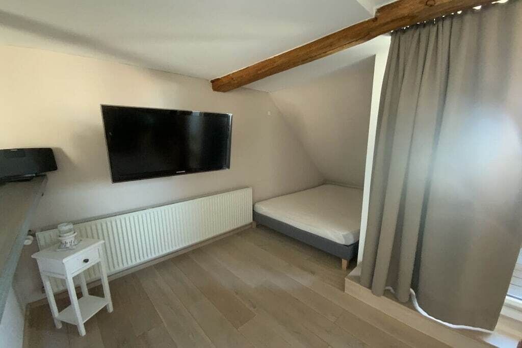 A small, single-room apartment with a bed, a television mounted on the wall, a radiator, and a small white table.