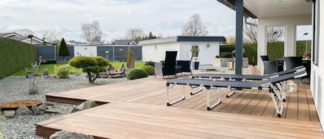 A wooden deck with outdoor furniture, a covered seating area, and a small building in the background.