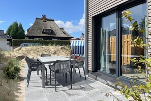 A patio with a table and chairs, a thatched-roof house in the background, and a wooden fence. - Holiday home with 4 bedrooms (Timmendorfer Strand)