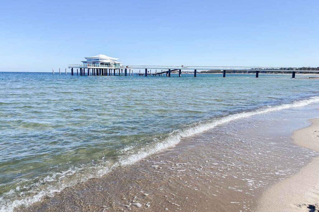 A beach with a pier extending into the water, a building on the pier, and a clear sky.