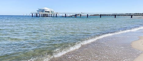 Una playa con un muelle que se adentra en el agua, un edificio sobre el muelle y un cielo despejado.