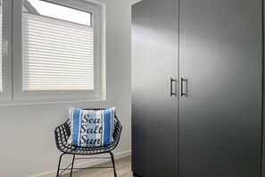 A room with a grey wardrobe, a window with blinds, and a chair with a pillow. - Holiday home with 4 bedrooms (Timmendorfer Strand)
