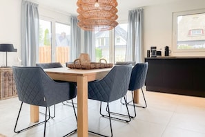 A dining area with a wooden table, four chairs, a woven light fixture, and a view of the outside through large windows. - Holiday home with 4 bedrooms (Timmendorfer Strand)