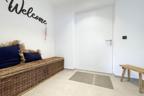 A clean, modern entryway with a white door, a wicker bench with cushions, a small wooden bench, and a welcome sign on the wall. - Holiday home with 4 bedrooms (Timmendorfer Strand)