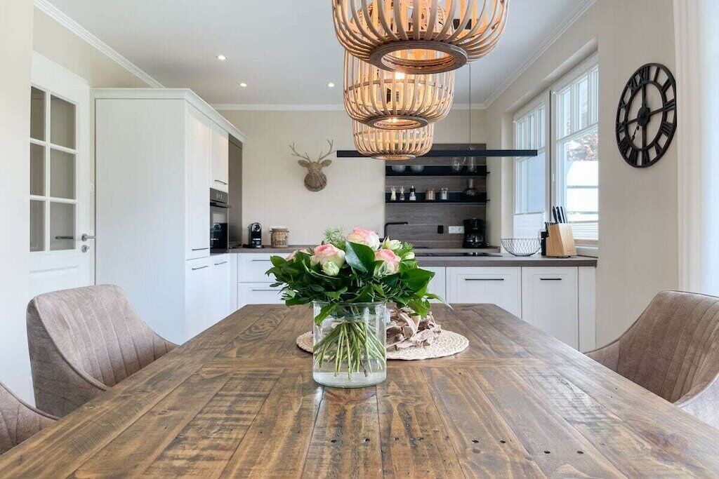 A dining area with a wooden table, chairs, a vase with flowers, a wall-mounted clock, and a kitchen in the background.