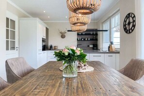 A dining area with a wooden table, chairs, a vase with flowers, a wall-mounted clock, and a kitchen in the background. - Holiday home with 4 bedrooms (Timmendorfer Strand)