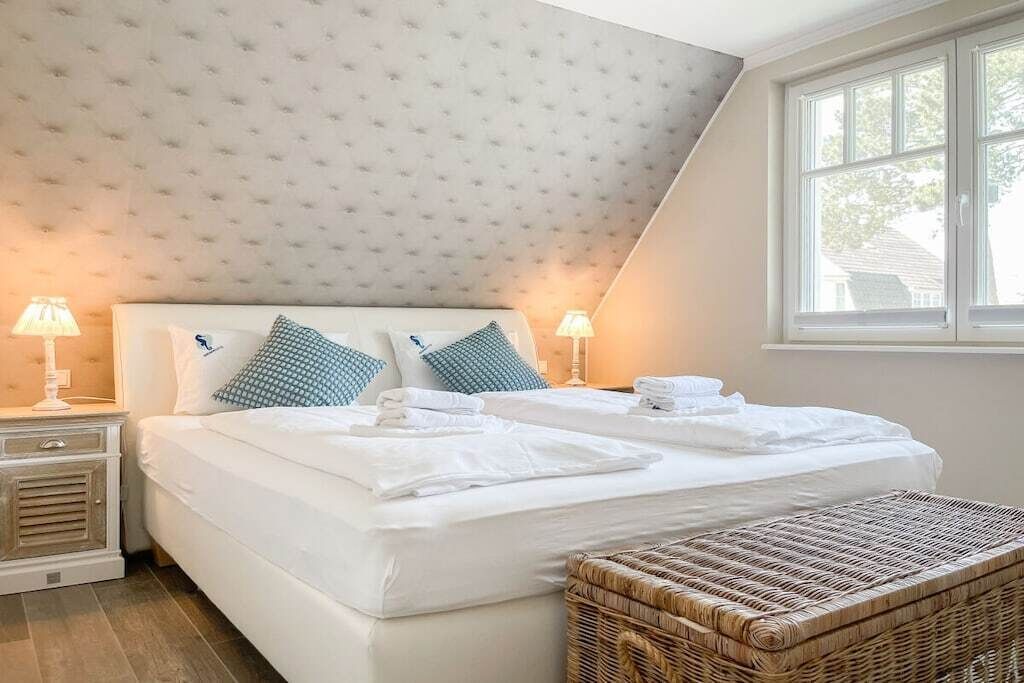 A bedroom with a large bed, two bedside lamps, a wicker bench, and a window with a view.