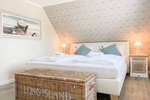A bedroom with a bed, two bedside lamps, a framed picture on the wall, and a wicker chest labeled 'LONG ISLAND'. - Holiday home with 4 bedrooms (Timmendorfer Strand)