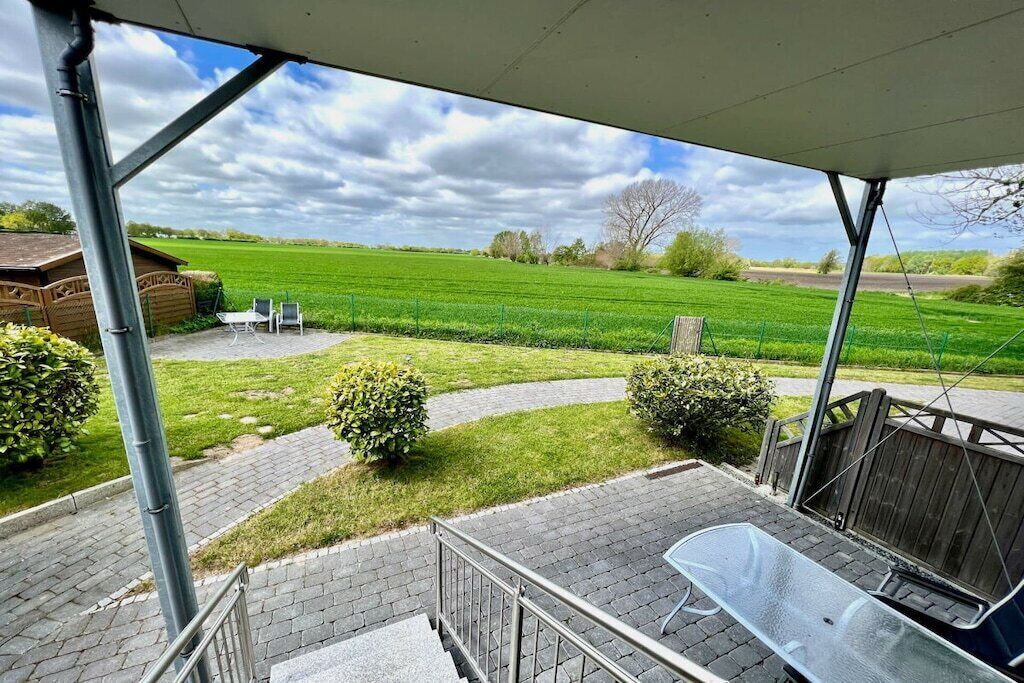 Ein überdachter Außenbereich mit Terrasse, Tisch und Stühlen mit Blick auf ein grünes Feld und eine Holzkonstruktion.