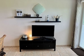 A modern living room with a flat-screen TV on a black stand, a small table with a lamp, and two black shelves with decorative items. - Holiday apartment in Dahme with swimming pool (Dahme)