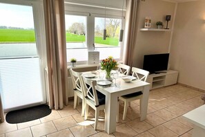 A dining area with a table set for four, a television on a stand, and a view of a green field through the window. - Holiday apartment in Dahme with swimming pool (Dahme)