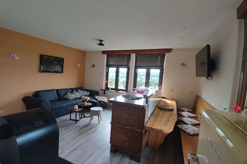 A living room with a sofa, a coffee table, a wooden bench, and a kitchen area with a counter and windows.