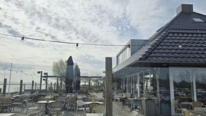 An outdoor seating area with tables and chairs, overlooking a body of water and a building with a dark roof.