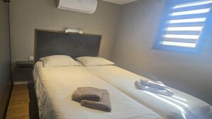 A single-bed room with a headboard, an air conditioning unit, a bedside table, a window with blinds, and a folded towel on the bed.