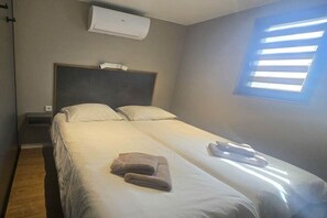 A single-bed room with a headboard, an air conditioning unit, a bedside table, a window with blinds, and a folded towel on the bed. - ||----------houseBoat Tranquility------|| (Offingawier)
