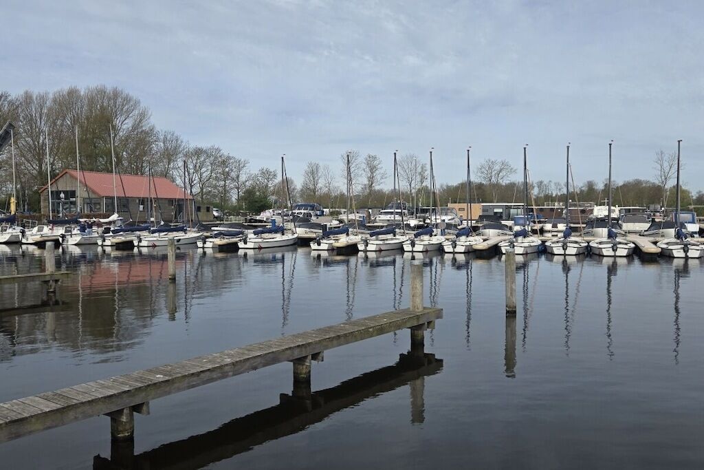 Un port de plaisance où sont amarrés de nombreux bateaux, une jetée en bois et, au fond, un bâtiment avec un toit rouge.