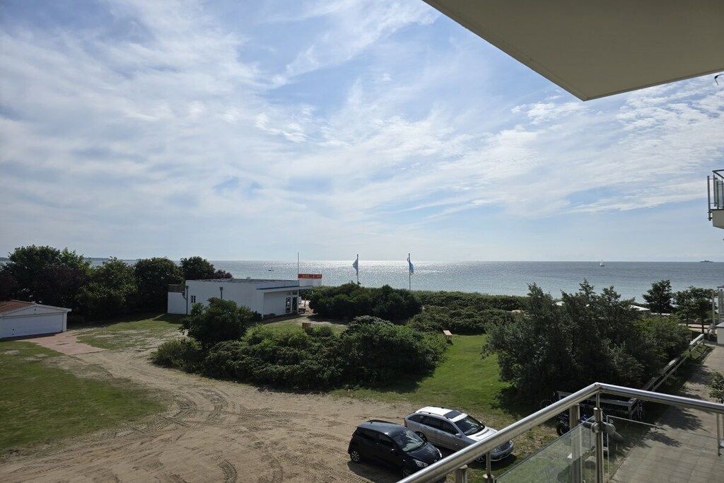 Uitzicht op de kust vanaf een balkon, met blauwe lucht, een gazon, geparkeerde auto's en een klein gebouw.
