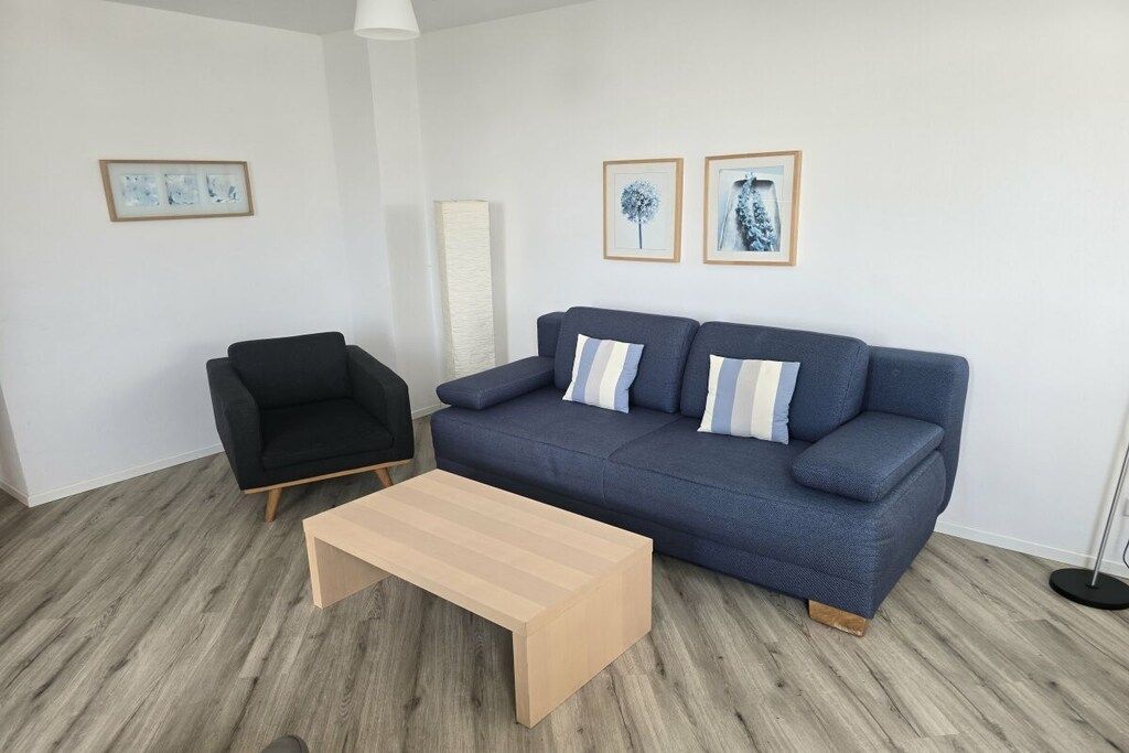 A living room with a blue sofa, a black armchair, and a wooden coffee table. There are two framed pictures on the wall.