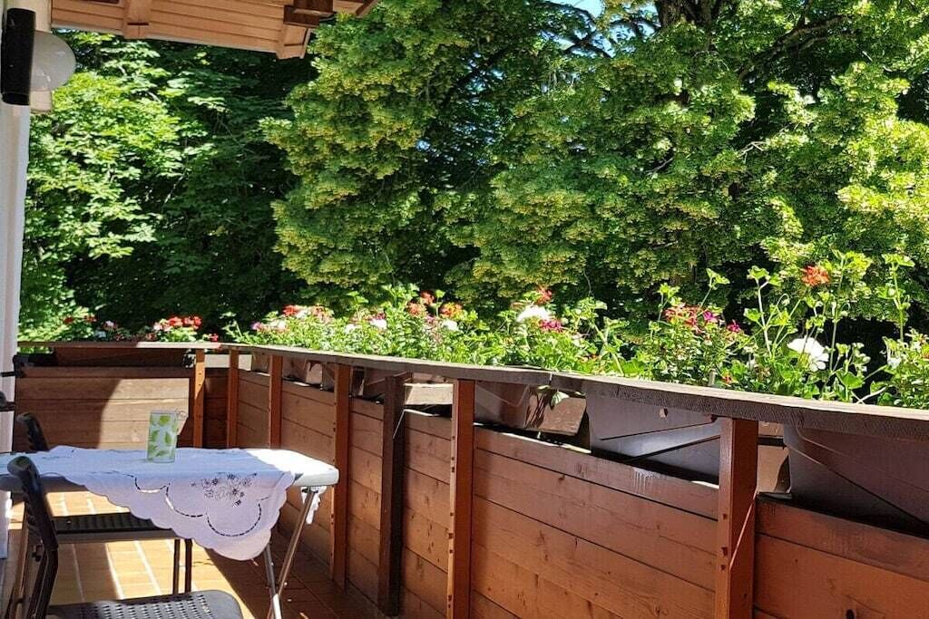 A wooden deck with a table and chairs, surrounded by lush greenery and flowering plants.
