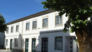 Un edificio blanco con varias ventanas y puertas, una calle adoquinada y un árbol en primer plano.