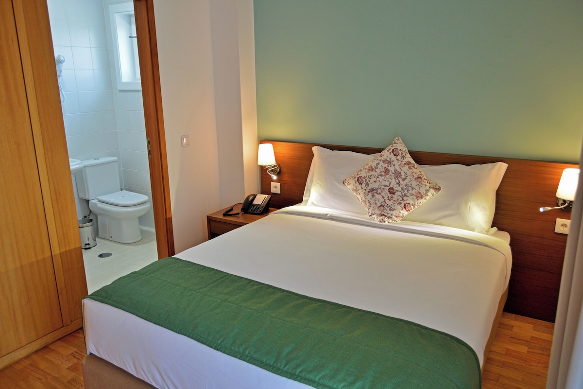 A hotel room with a bed, a bedside table with a lamp, a telephone, and a floral pillow.