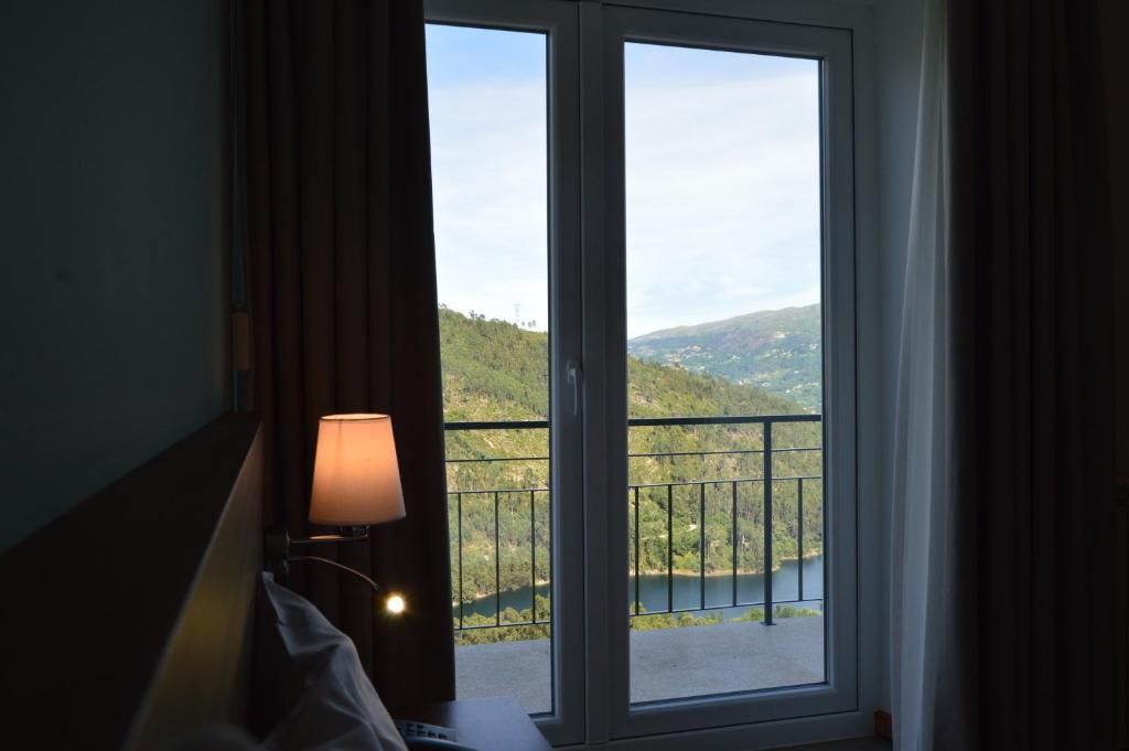 Una habitación con vista a un lago y colinas, una cama, una lámpara y una ventana con cortinas.