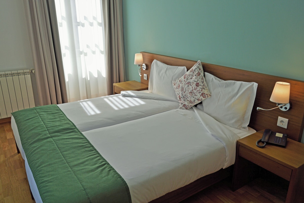 A double bed with a green bedspread, two bedside lamps, a floral pillow, and a telephone on a wooden nightstand.