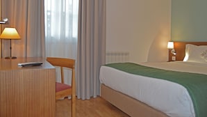 Habitación de hotel con una cama, un escritorio con lámpara, una silla y una ventana con cortinas.