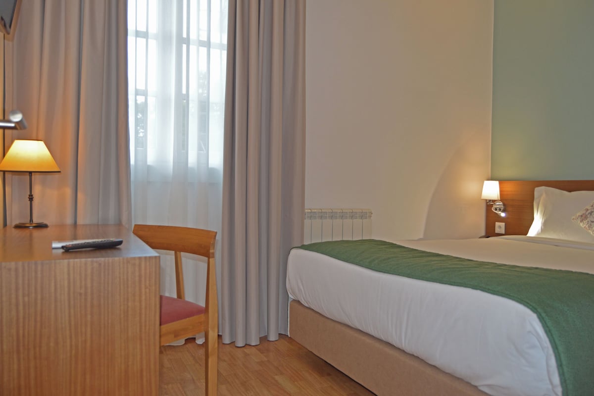 A hotel room with a bed, a desk with a lamp, a chair, and a window with curtains.