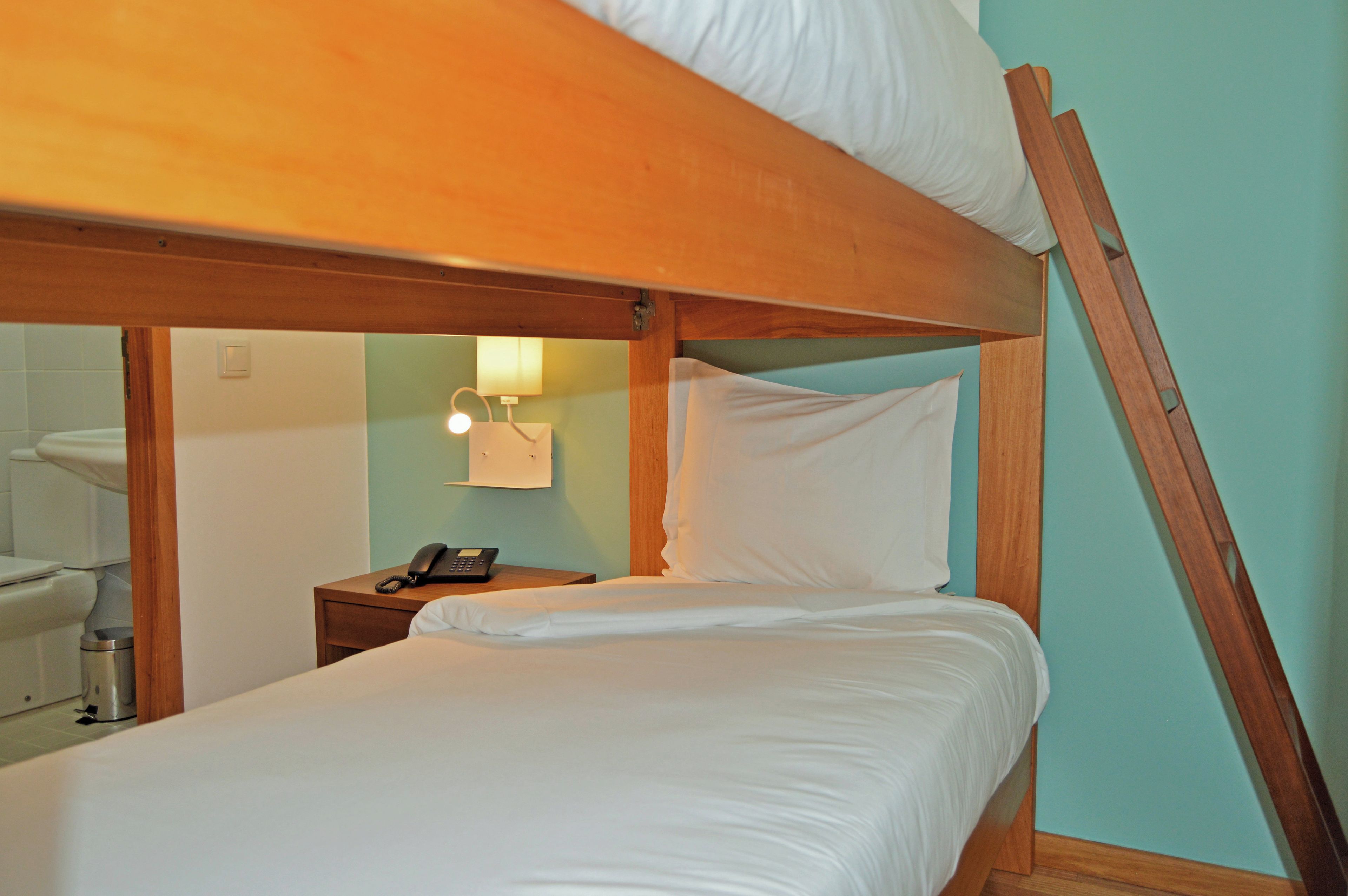 A bunk bed with a wooden frame, a bedside table with a phone, and a wall-mounted light fixture.