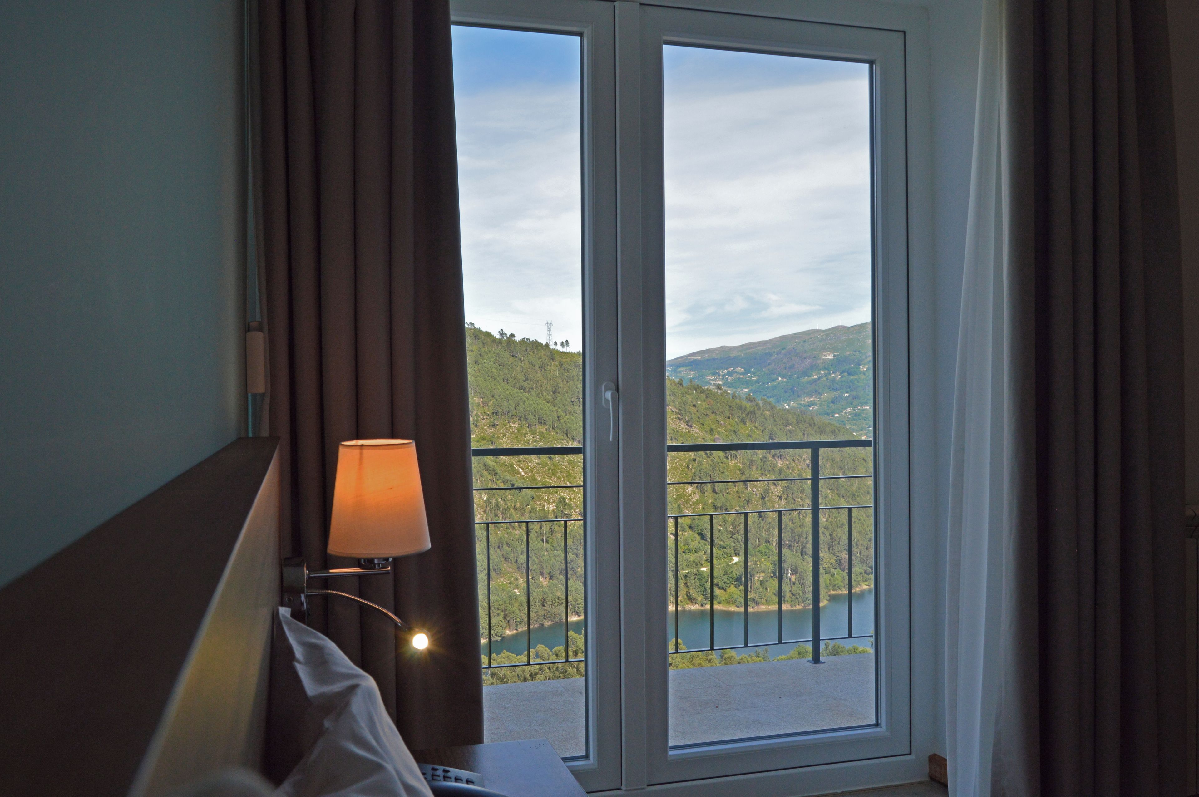 Habitación de hotel con cama, mesita de noche, lámpara y vista a un lago y colinas a través de un ventanal.