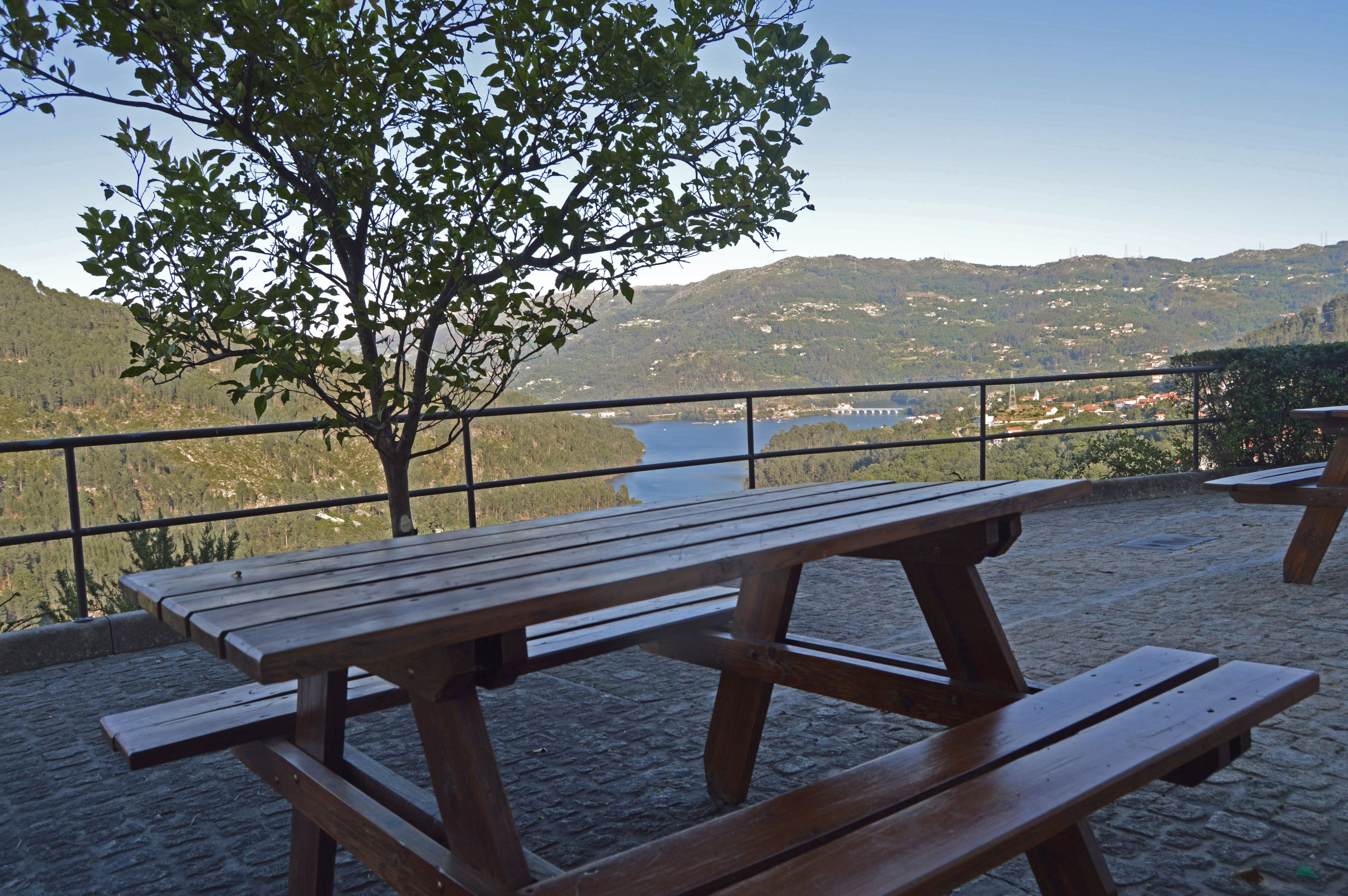 Una mesa de picnic de madera con bancos, una baranda metálica y una vista de un lago y colinas al fondo.