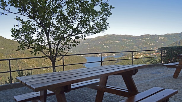 Una mesa de picnic de madera con bancos, una baranda metálica y una vista de un lago y colinas al fondo.