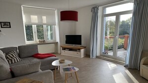 Ein modernes Wohnzimmer mit einem grauen Sofa, einem kleinen weißen Tisch, einer roten Pendelleuchte, einem Fernseher auf einem Holzständer und einem großen Fenster mit Vorhängen.