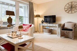 Een gemeubileerde woonkamer met een televisie, een tafel met een modelhuis, een rieten stoel en een klok aan de muur.